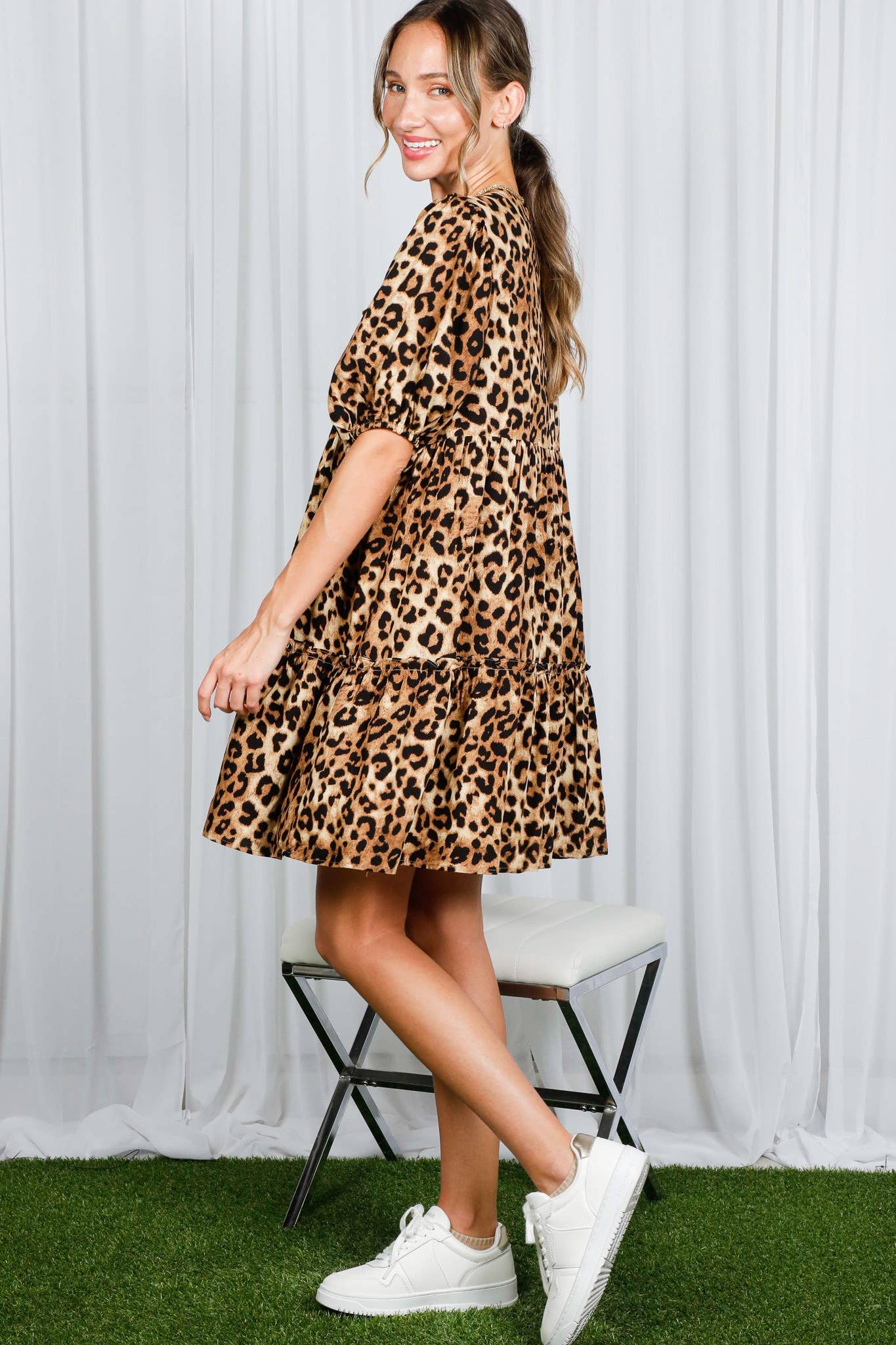 Woman wearing a leopard print dress with white sneakers against a white curtain background