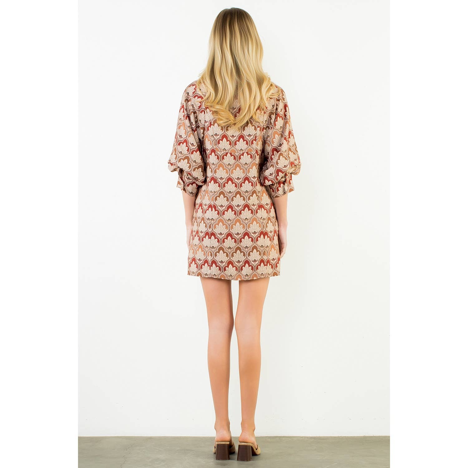 A woman viewed from behind wearing a short puff-sleeve dress with a beige, brown, and rust-red geometric pattern, standing against a plain white wall.