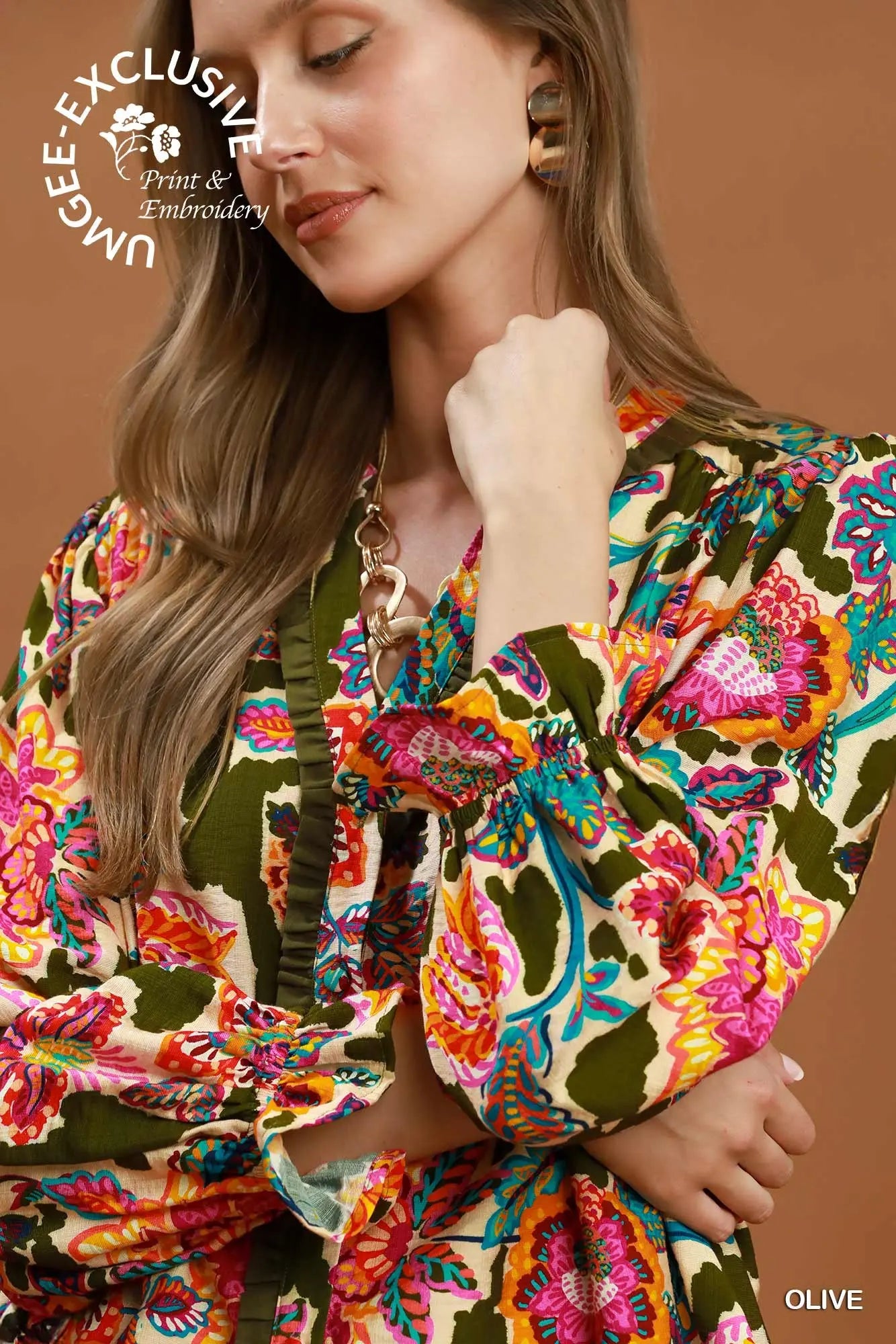 A close-up of a woman wearing a vibrant, multicolored floral print blouse with olive green ruffled trim and puff sleeves, set against a solid warm brown background.