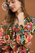 A close-up of a woman wearing a vibrant, multicolored floral print blouse with olive green ruffled trim and puff sleeves, set against a solid warm brown background.