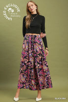 A woman modeling a black long-sleeve crop top and a high-waisted, wide-leg midi skirt with a vibrant paisley print in pink, purple, blue, and yellow on a black base. She is standing against a solid olive green studio background.