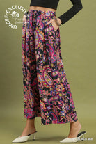 A model wears high-waisted, wide-leg cropped pants with a vibrant multicolor paisley print on a black base, styled with a black long-sleeve crop top and white pointed-toe mule heels against a solid olive green background.