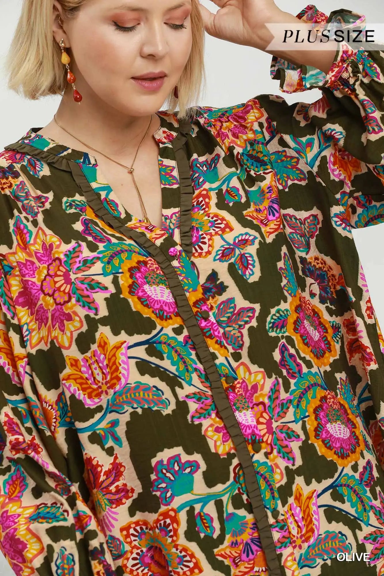 A close-up of a woman wearing a loose, long-sleeve blouse with a vibrant multicolor floral and paisley print on a beige base, featuring dark olive green ruffle trim along the front and neckline. The brand name 'PLUS SIZE' is visible in the top right corner.
