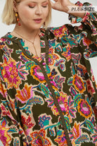 A close-up of a woman wearing a loose, long-sleeve blouse with a vibrant multicolor floral and paisley print on a beige base, featuring dark olive green ruffle trim along the front and neckline. The brand name 'PLUS SIZE' is visible in the top right corner.