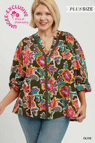 A woman wearing a short-sleeve button-front blouse with a colorful floral and paisley print over an olive base, paired with blue jeans. The image includes text overlays reading 'PLUS SIZE', 'OLIVE', and a circular 'UNIQUELY EXCLUSIVE Print & Embroidery' badge.