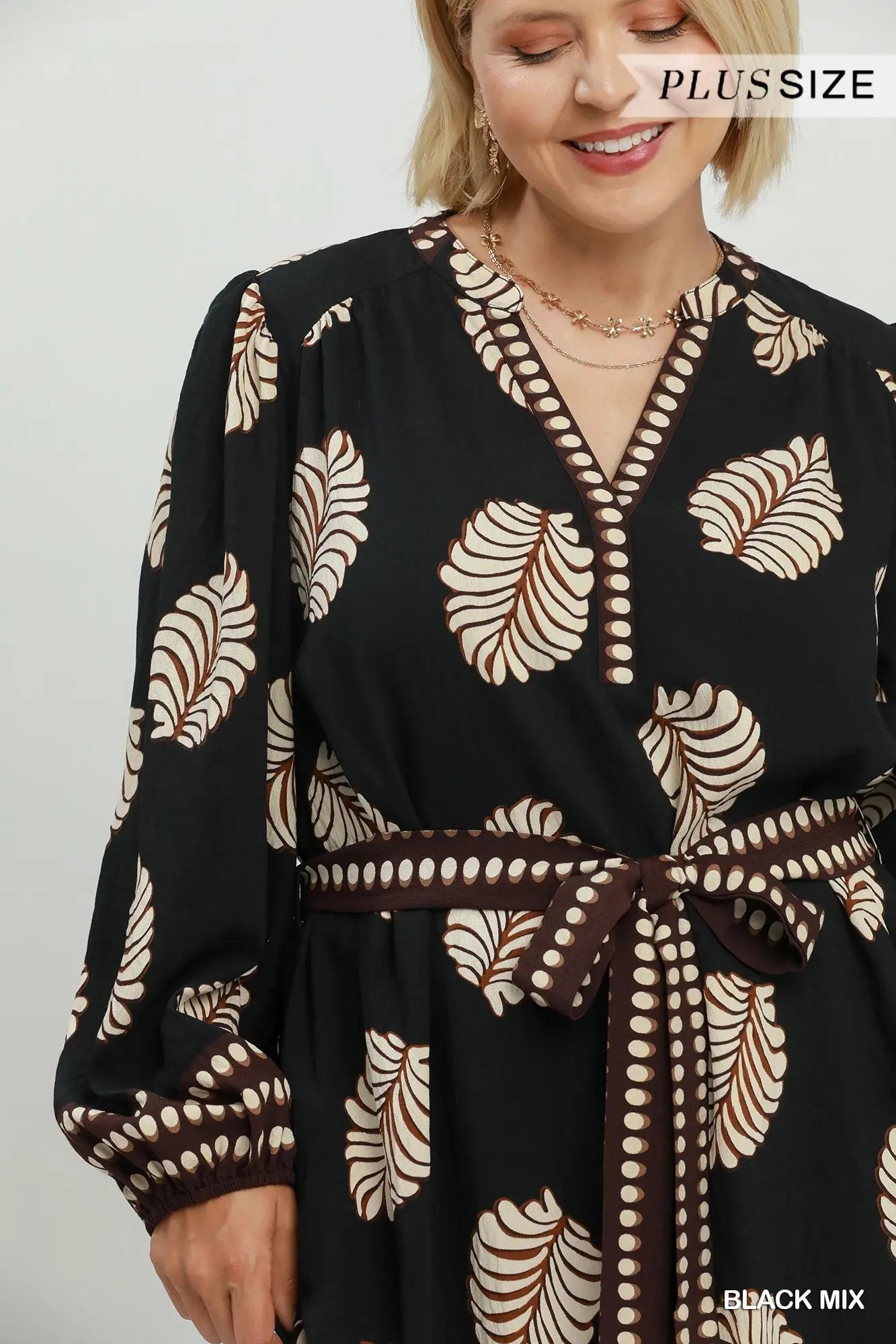A woman wearing a black long-sleeve wrap dress with a beige and brown leaf pattern, a matching brown belt, and a V-neckline. The brand name 'PLUSSIZE' is visible in the top right corner.