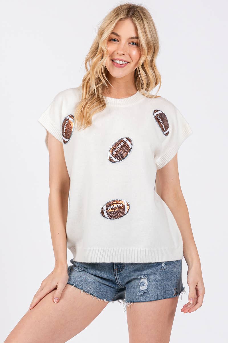 A woman modeling a white short-sleeve knit top with brown football graphics, paired with distressed denim shorts, against a light gray studio background.