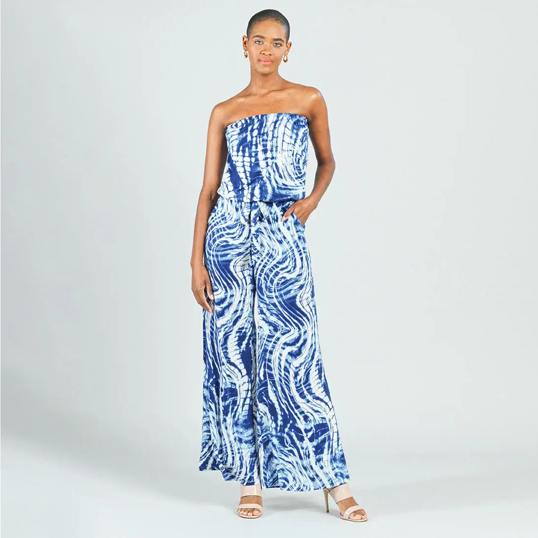 A woman modeling a strapless blue and white tie-dye wide-leg jumpsuit against a light gray studio background.