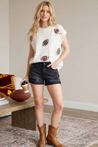 A woman stands indoors wearing a cream knit sweater with football patches, black denim shorts, and brown cowboy boots, holding a coffee cup beside a wooden bench with a football and blanket.