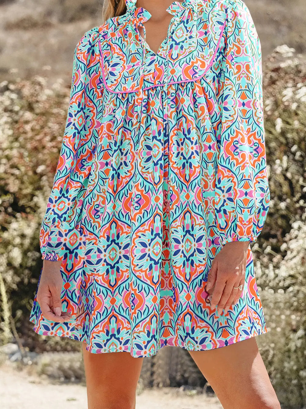 A woman wearing a long-sleeve bohemian-style mini dress with a vibrant multicolor paisley and floral pattern in turquoise, orange, pink, and purple, set against a blurred outdoor background.