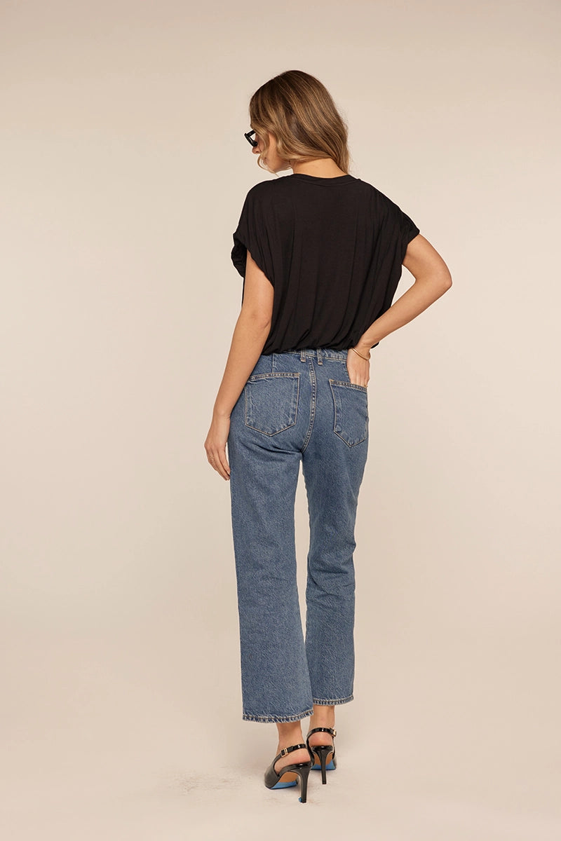A woman is shown from behind wearing blue cropped jeans, a black short-sleeve top, and black high-heeled sandals against a plain beige studio background.