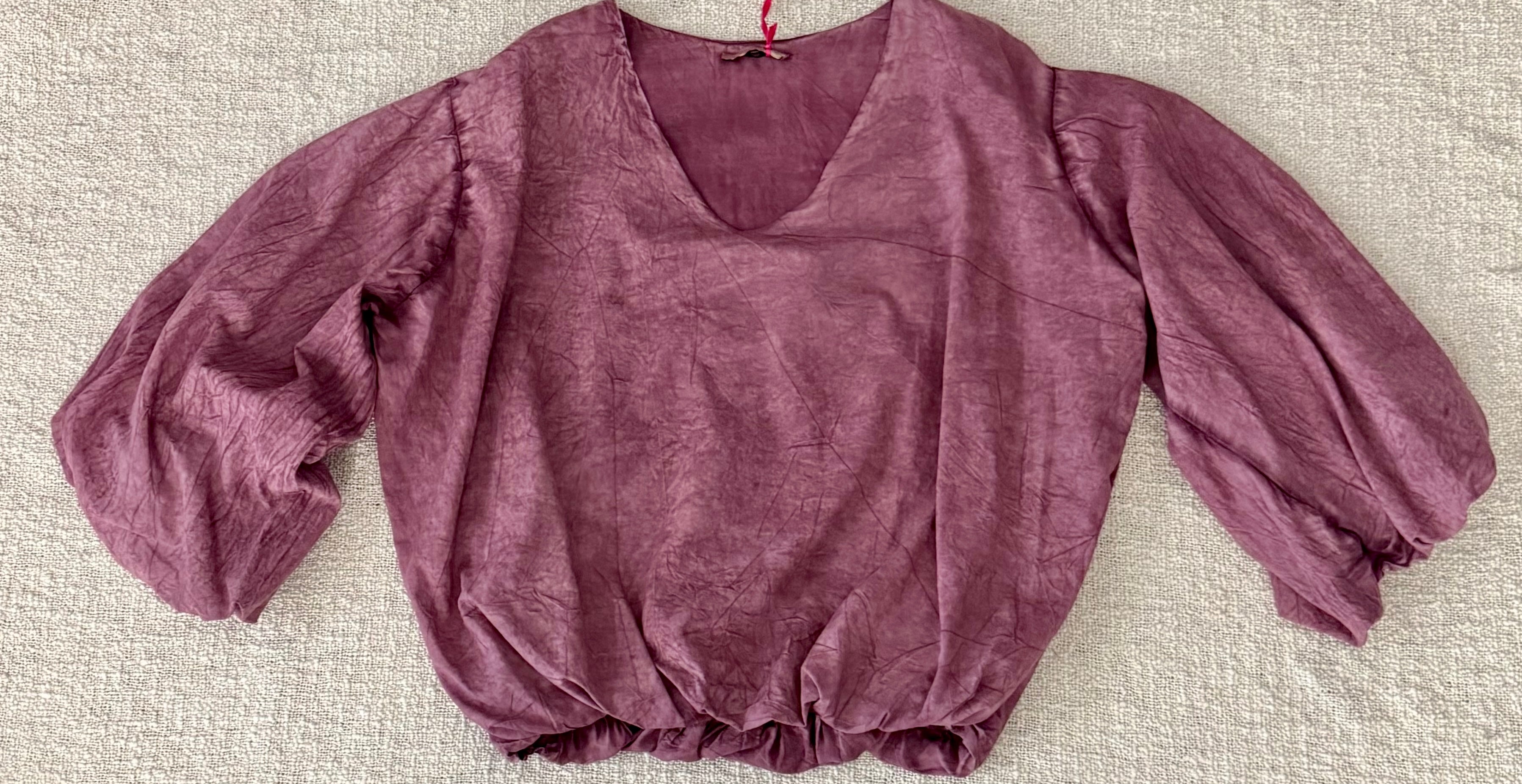 A dusty mauve-purple crinkled cotton blouse with a V-neckline and voluminous elbow-length puff sleeves, laid flat on a textured beige fabric background.