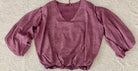 A dusty mauve-purple crinkled cotton blouse with a V-neckline and voluminous elbow-length puff sleeves, laid flat on a textured beige fabric background.