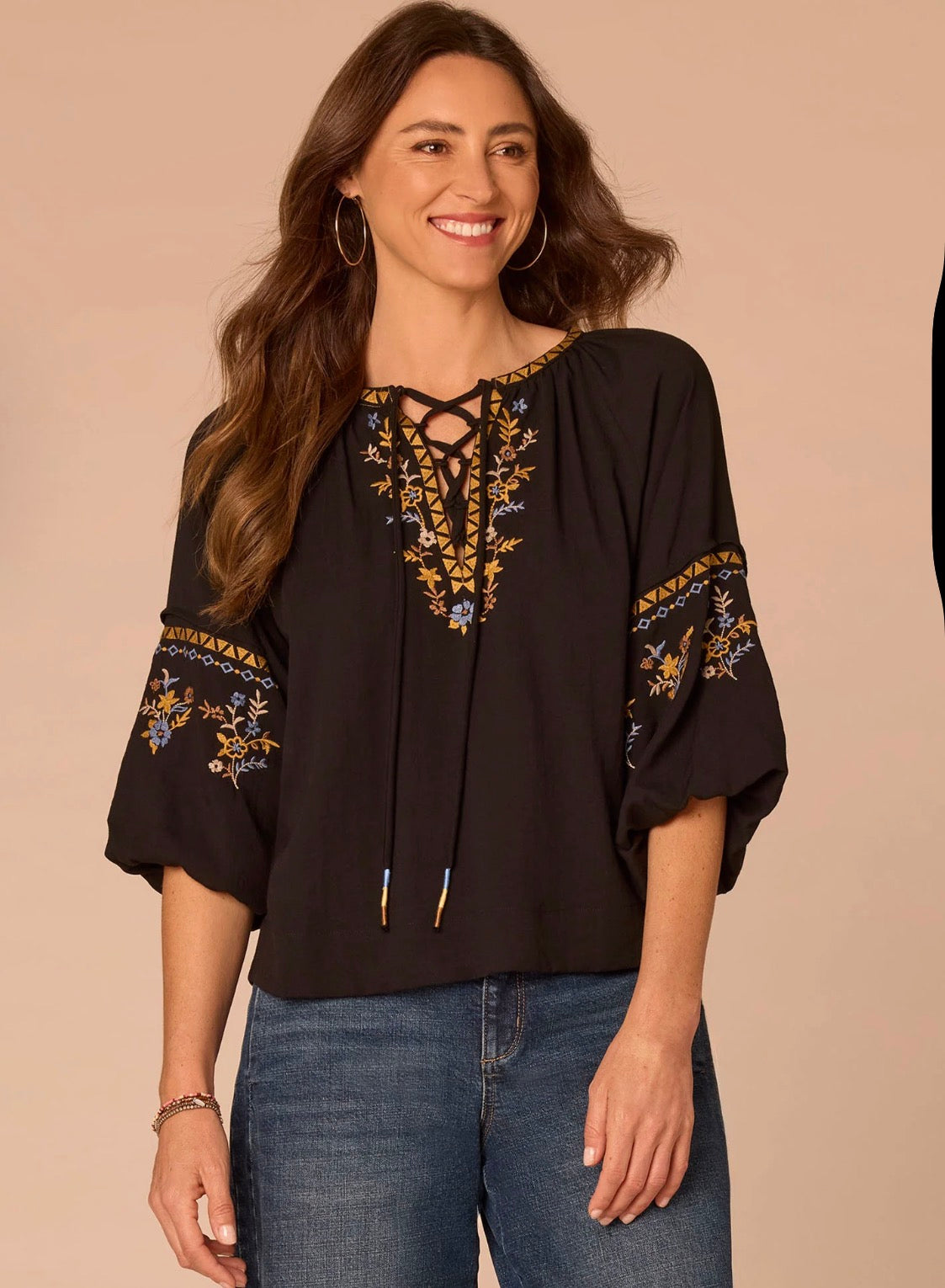 A woman wearing a black bohemian-style blouse with gold, blue, and white floral embroidery on the neckline and sleeves, paired with blue jeans, set against a solid beige background.