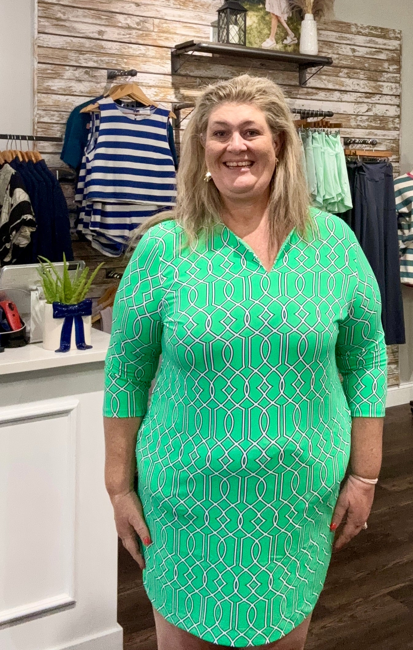 A woman modeling a mint green V-neck dress with a white geometric pattern, standing inside a clothing boutique.