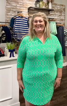 A woman modeling a mint green V-neck dress with a white geometric pattern, standing inside a clothing boutique.