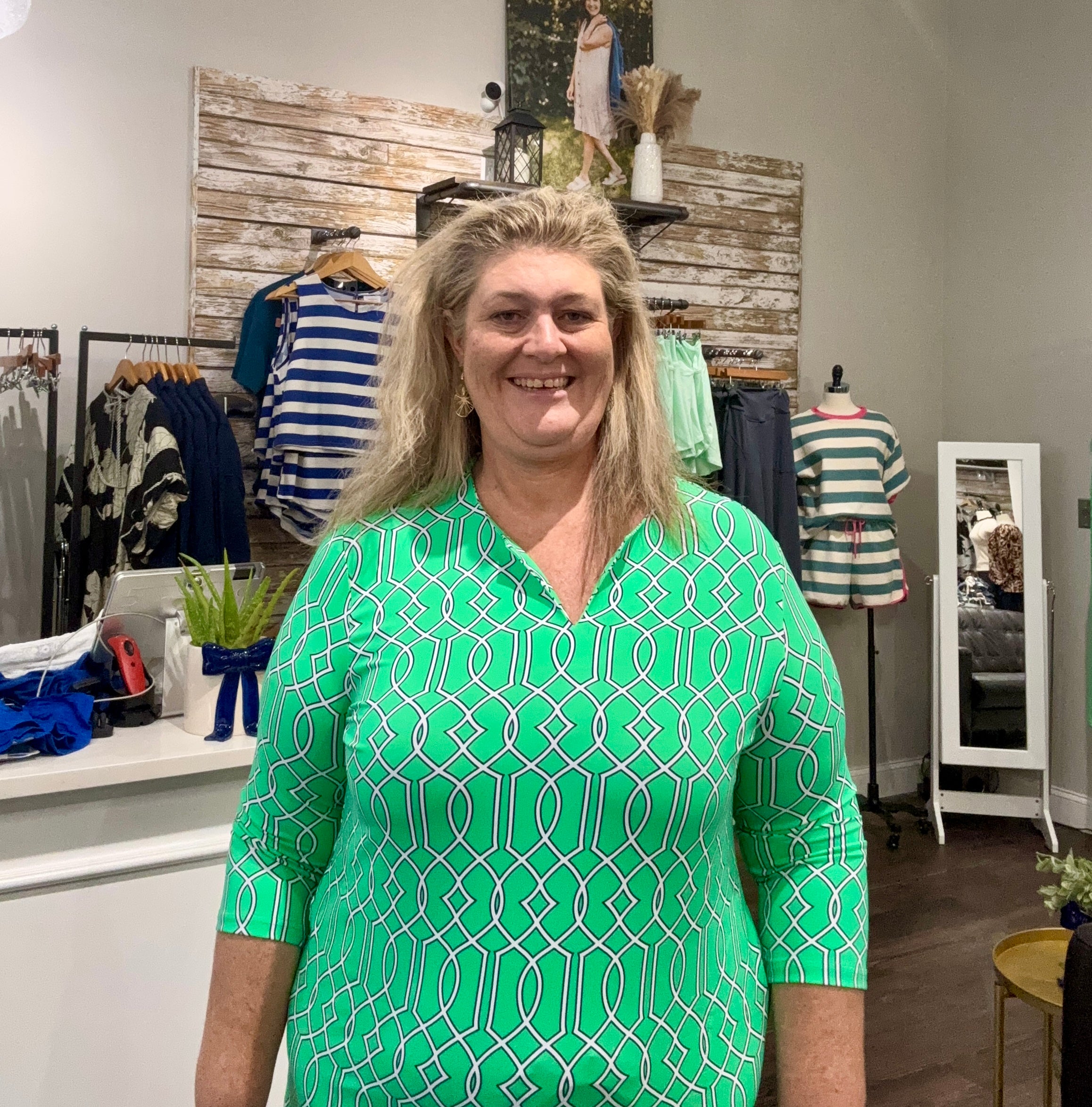 A smiling woman with blonde hair wearing a bright green and white geometric patterned short-sleeve top, standing inside a clothing boutique.