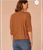 A woman shown from the back wearing a rust-brown cable-knit cardigan with three-quarter sleeves and blue jeans, set against a solid beige background.
