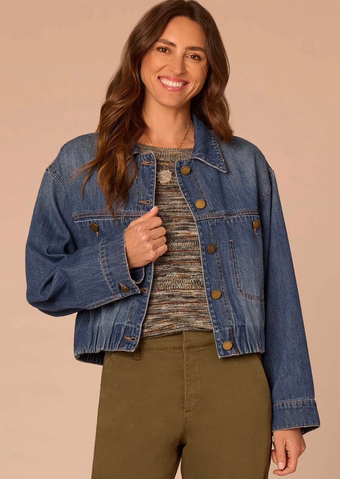 A woman wearing a cropped blue denim jacket over a textured knit top and olive green pants, standing against a solid beige background.