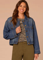 A woman wearing a cropped blue denim jacket over a textured knit top and olive green pants, standing against a solid beige background.