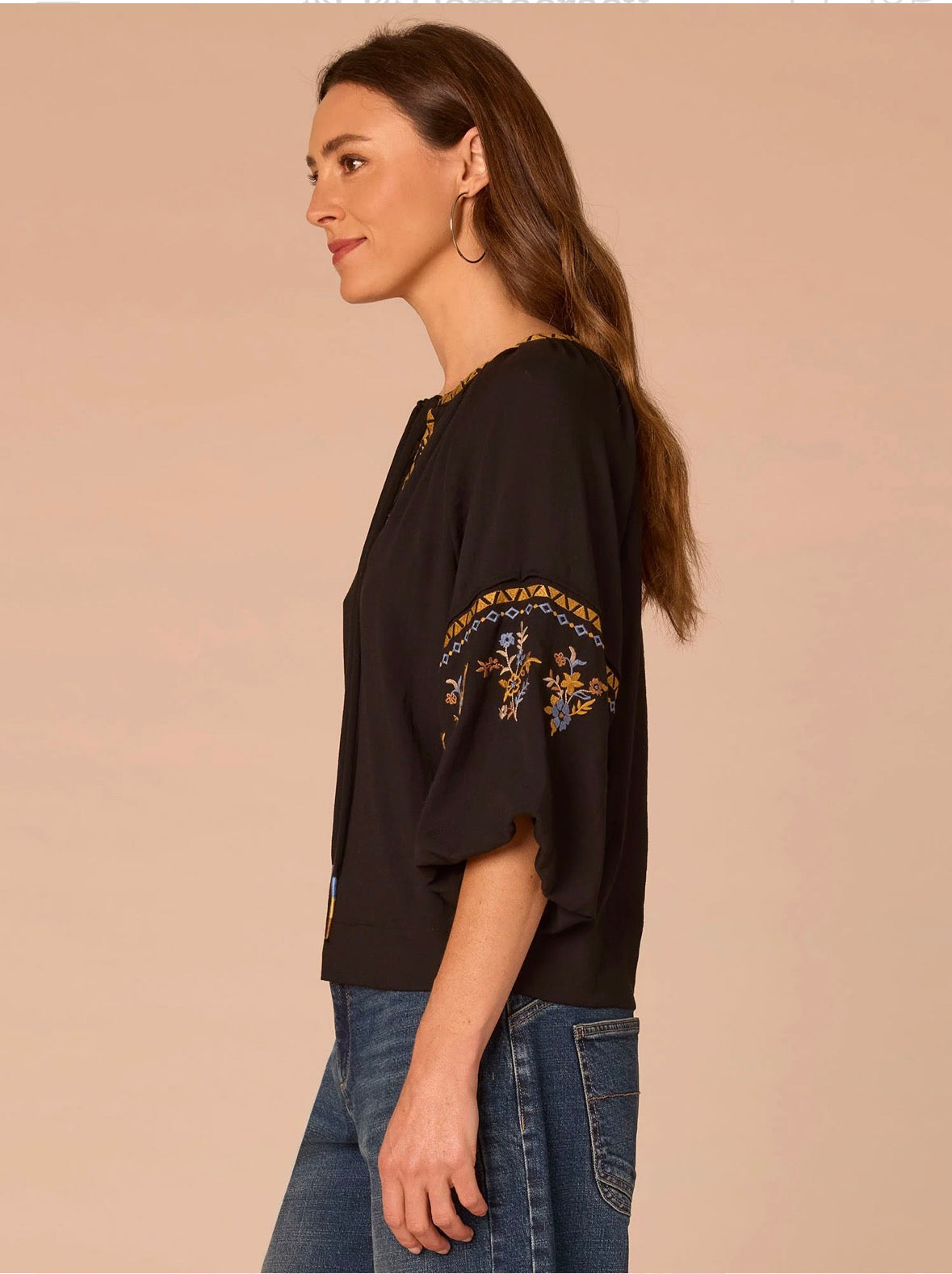 A woman shown in side profile wearing a black short-sleeve top with gold and blue embroidery on the sleeve, paired with blue jeans, against a solid beige background.