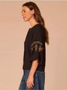 A woman shown in side profile wearing a black short-sleeve top with gold and blue embroidery on the sleeve, paired with blue jeans, against a solid beige background.