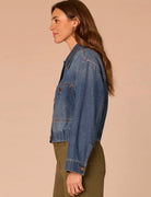 A woman shown in side profile wearing a cropped blue denim jacket over an olive green top, set against a solid warm beige studio background.