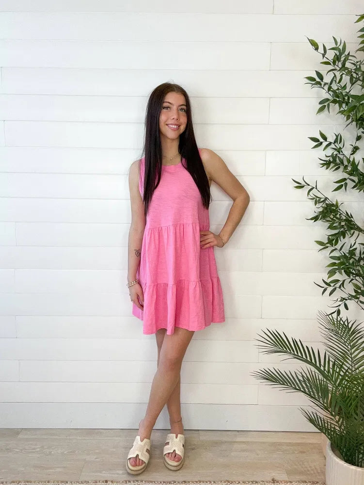 Model wearing tiered, candy, pink, mini dress with neutral wedges against a white wall