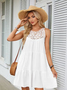 A woman wearing a white sleeveless tiered dress with a crochet lace neckline, a wide-brimmed straw hat, and a woven brown shoulder bag, standing in front of a white shuttered building.