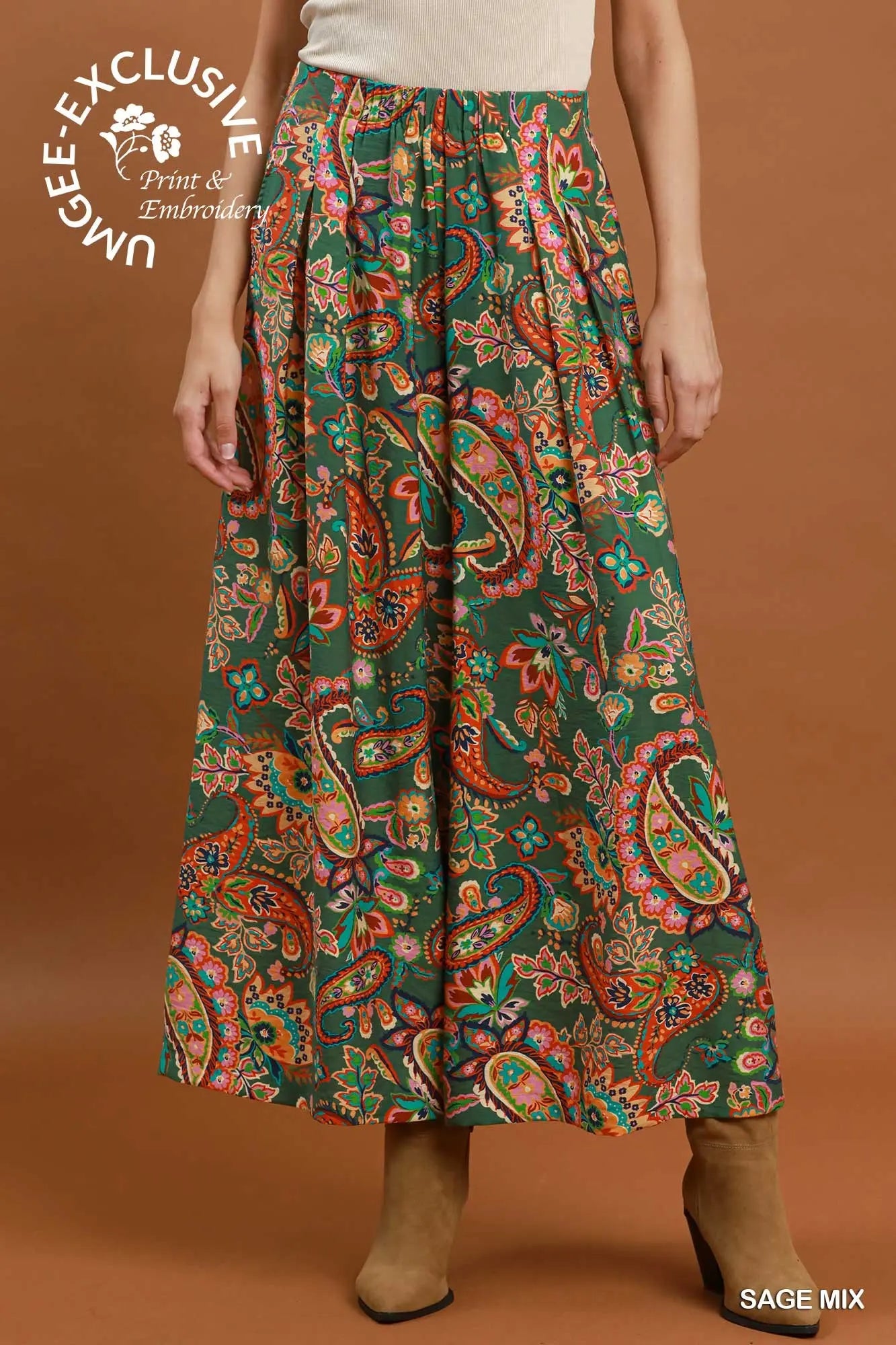 A model is shown from the waist down wearing a high-waisted, wide-leg maxi skirt with a vibrant paisley pattern in green, orange, red, and yellow. The background is a solid warm brown studio backdrop.