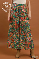 A model is shown from the waist down wearing a high-waisted, wide-leg maxi skirt with a vibrant paisley pattern in green, orange, red, and yellow. The background is a solid warm brown studio backdrop.