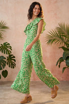 A woman modeling a green and white floral print one-shoulder jumpsuit with wide-leg pants, styled with tan wedge sandals against a textured pink wall with tropical plants.