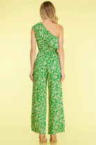 A woman viewed from the back wearing a bright green one-shoulder jumpsuit with a white floral print, set against a solid pale yellow background.