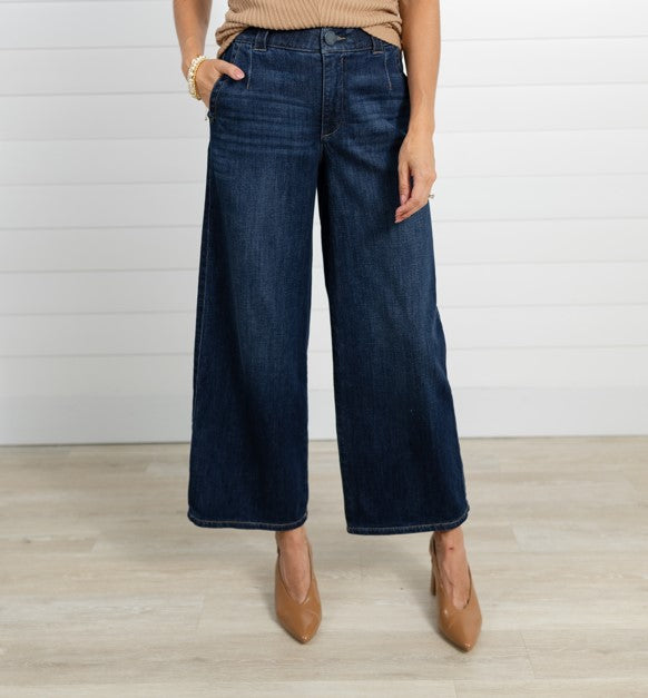 A model wearing dark blue wide-leg cropped jeans, paired with a beige ribbed top and tan pointed-toe heels, standing against a white shiplap wall and light wood floor.
