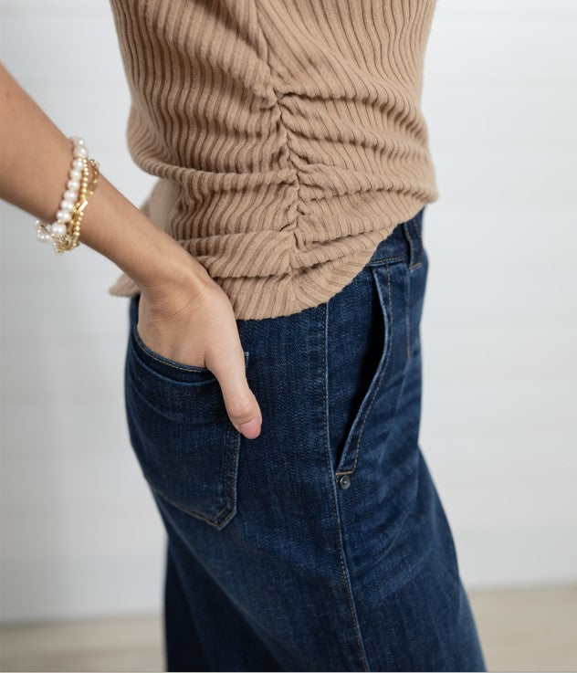 A close-up of a person wearing a beige ruched ribbed knit top and dark blue denim jeans, with one hand in a pocket and pearl-and-gold bracelets on the wrist.