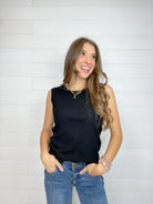 A woman wearing a sleeveless black top and blue jeans, standing against a white shiplap wall.