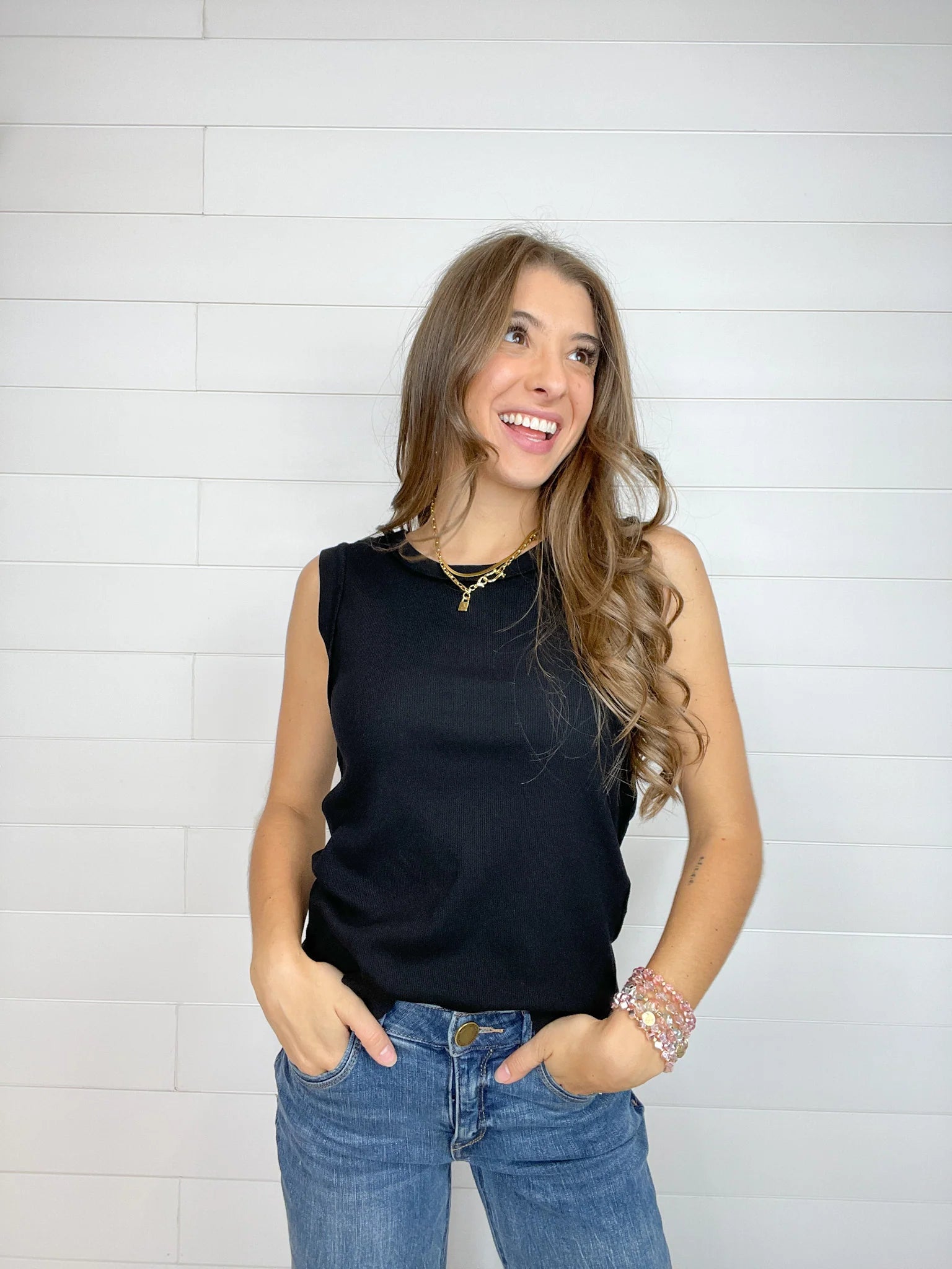 A woman wearing a sleeveless black top and blue jeans, standing against a white shiplap wall.