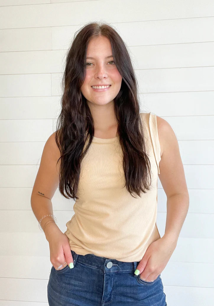 A woman with long dark brown hair smiling, wearing a light beige sleeveless tank top and blue jeans, standing against a white shiplap wall.