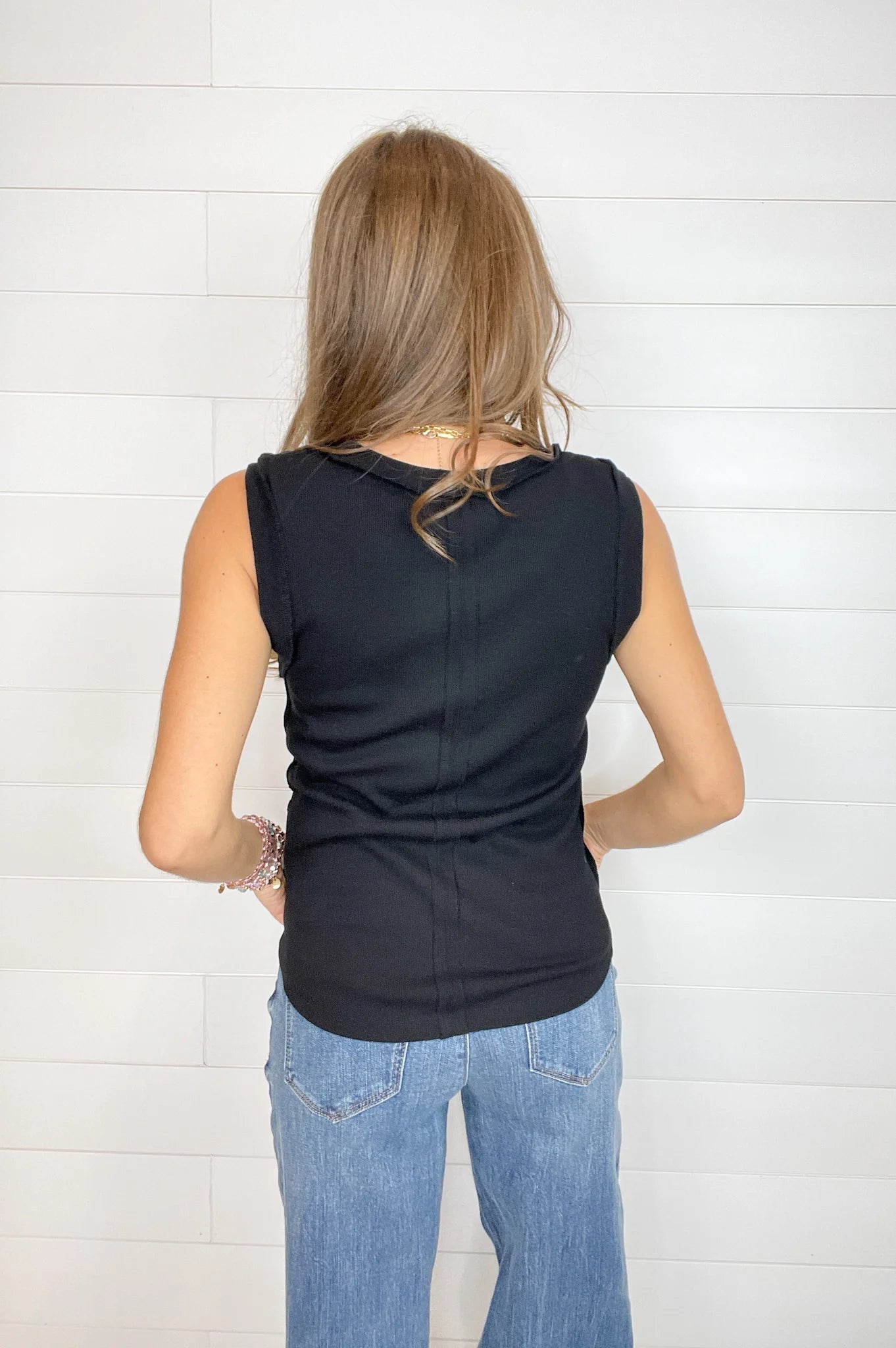 A woman viewed from behind wearing a black sleeveless top and light blue jeans, standing against a white shiplap wall.