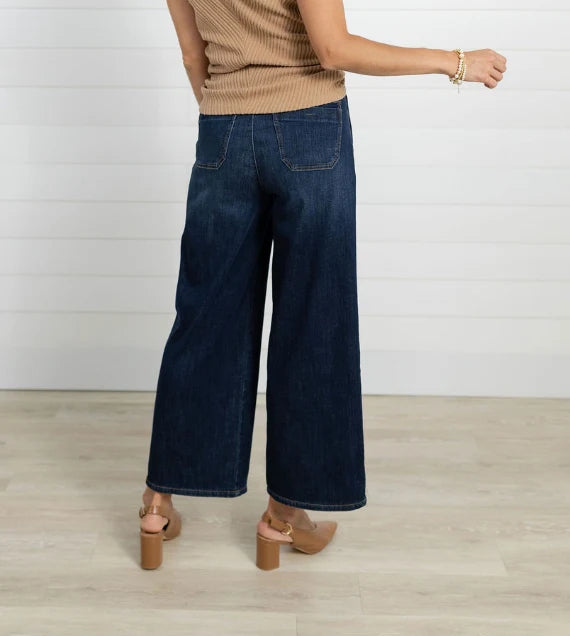 A person shown from the back wearing dark blue wide-leg denim jeans, a beige ribbed knit top, and tan block-heel sandals against a white paneled wall and light wood floor.