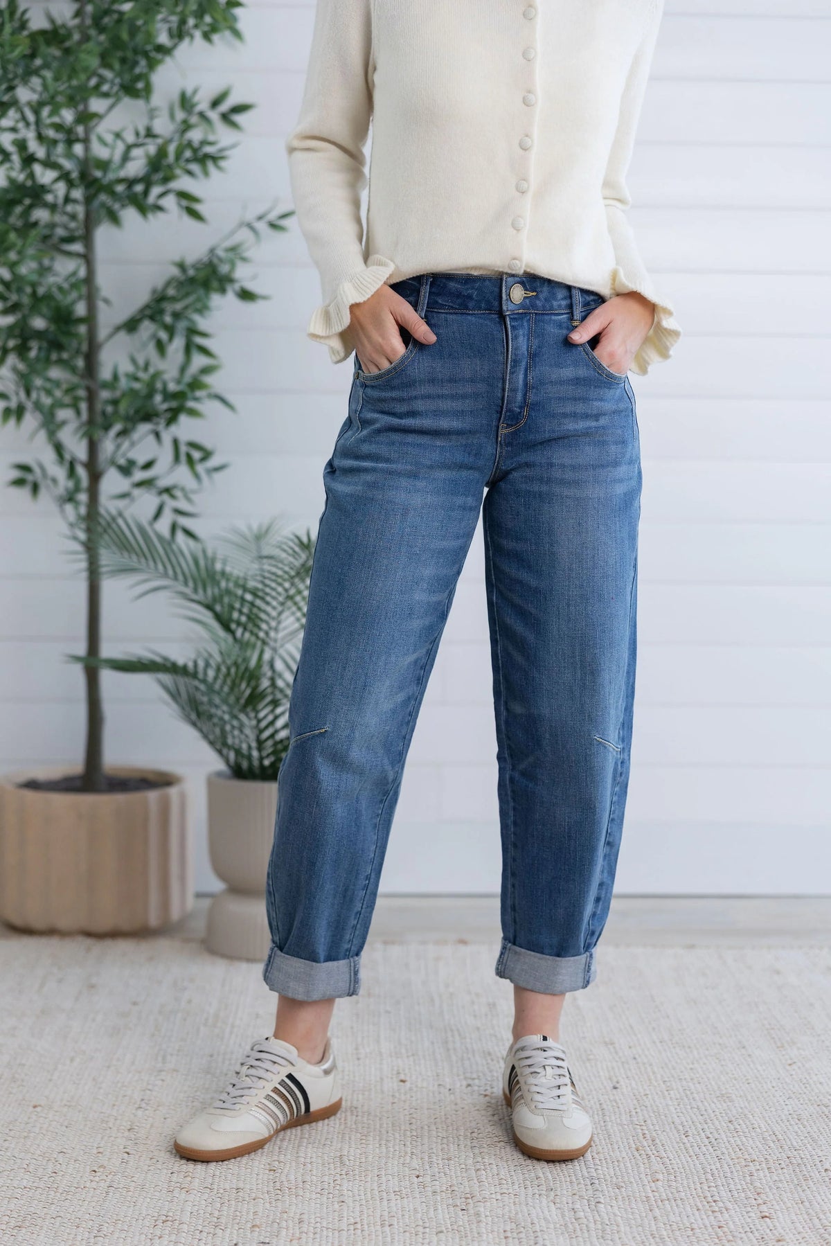 A person wearing blue cuffed jeans, a cream button-up sweater, and white sneakers, standing in front of a white paneled wall with potted plants.