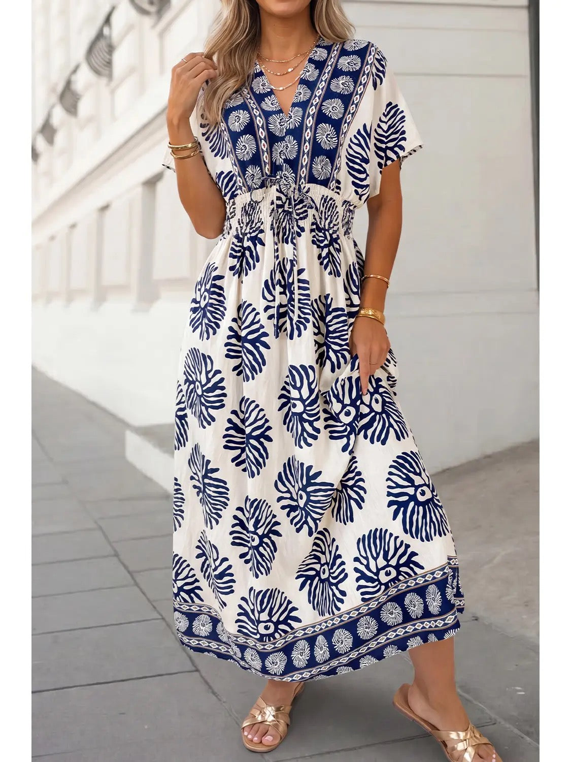 A woman wearing a white maxi dress with a navy blue tropical leaf and border print, standing outdoors on a paved walkway.
