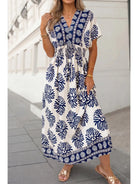A woman wearing a white maxi dress with a navy blue tropical leaf and border print, standing outdoors on a paved walkway.