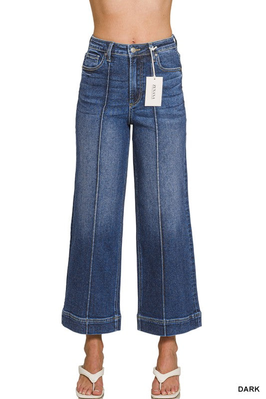 A model is wearing a pair of dark blue wide-leg cropped jeans with vertical seam detailing down the front. The jeans are shown from the waist down, with a white hangtag attached to the waistband and white slide sandals on the feet, against a plain white background.