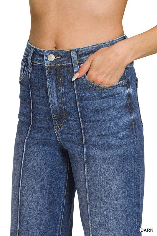 Close-up of a person wearing dark blue denim jeans, showing the front waistband, button, belt loops, front pockets, and a hand in the side pocket against a white background.