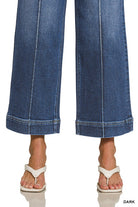 Close-up of a person wearing blue wide-leg cropped jeans with rolled hems and white thong sandals, shown against a white background with a small 'DARK' text label in the corner.
