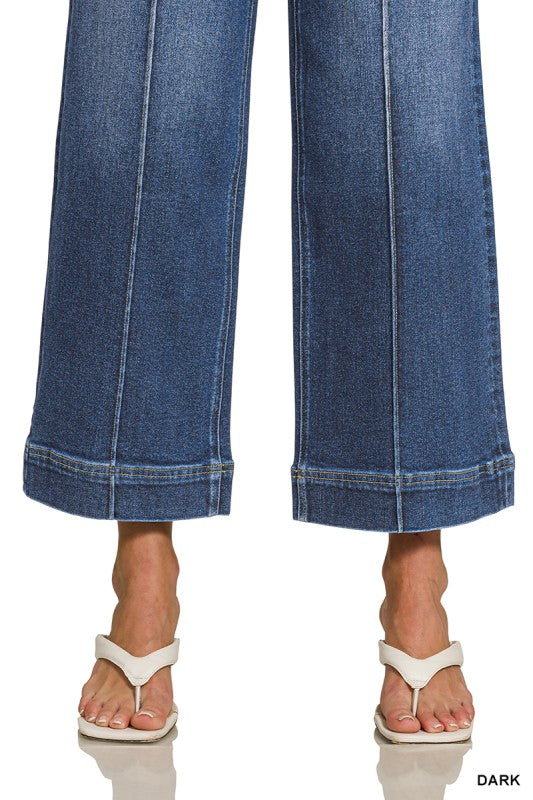 Close-up of a person wearing blue wide-leg cropped jeans with rolled hems and white thong sandals, shown against a white background with a small 'DARK' text label in the corner.