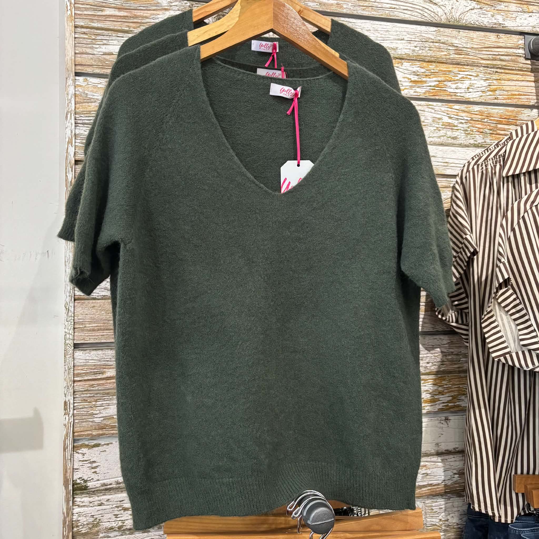 A dark olive green short-sleeve V-neck knit sweater hanging on a wooden hanger against a rustic whitewashed wood plank wall, with another matching sweater partially visible behind it and a striped shirt on the right.