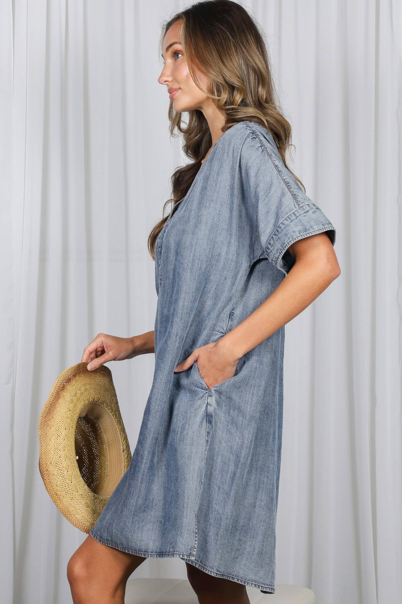 Woman wearing a denim dress holding a hat against a white curtain background