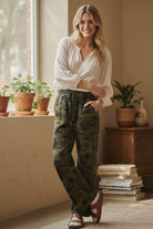 Woman wearing a white blouse and camouflage pants standing in a room with plants and books.